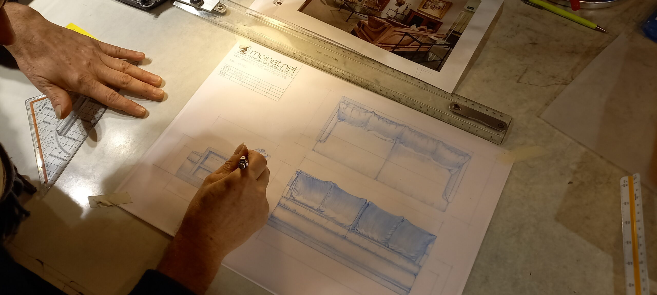Main en train de dessiner un canapé sur une feuille de papier calque avec des outils de dessin à côté.